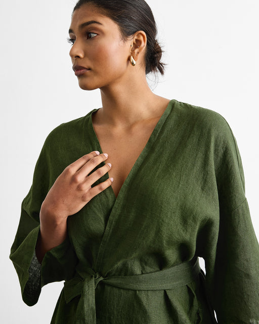 A woman with dark hair tied back wears the Bed Threads Cabana Short Robe in Olive, belted at the waist. She looks to the side with a neutral expression, one hand on her chest, gold hoop earrings accenting her look against a white background.