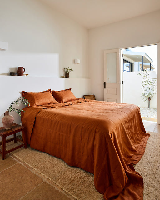 A cozy bedroom features Bed Threads' Rust Quilted Pillowcases, a burnt orange bedspread, a wooden side table with a vase, neutral walls, a natural fiber rug, and an open door to a bright outdoor plant-filled space.