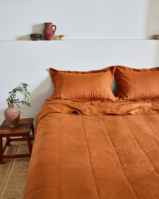 A neatly made bed features Bed Threads' Rust Quilted Pillowcases (Set of Two) in French flax linen, beside a small wooden table with a vase of greenery. Ceramic pots and bowls are arranged on a white shelf above the bed.