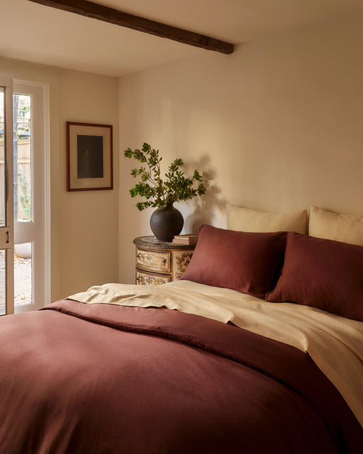 A cozy bedroom with cream walls, a burgundy and beige bed, and Bed Threads’ Crème 100% French Flax Linen European Pillowcases (Set of Two) propped against the headboard, sunlight streaming through glass doors.