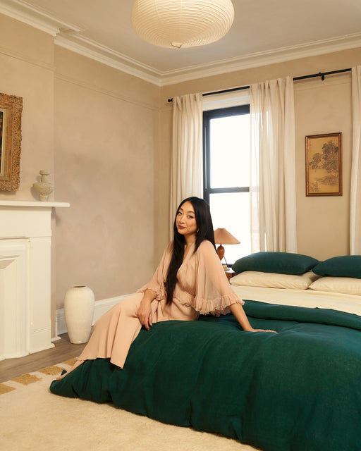 A woman in a light peach outfit sits on the edge of a bed with a Bed Threads Forest 100% French Flax Linen Duvet Cover in a softly lit, neutral-toned bedroom featuring large windows, cream curtains, wall art, and decorative vases.