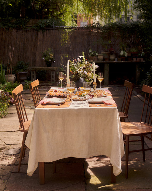 A sunlit garden patio table for four features a cream tablecloth, floral centerpiece, candles, wooden chairs, and Bed Threads' Pink Clay 100% French Flax Linen Napkins (Set of Four), plus plates of food and wine glasses among lush greenery.