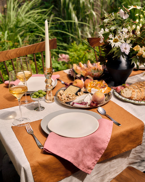 A table set outdoors with plates, Bed Threads Pink Clay 100% French Flax Linen Napkins (Set of Four), wine glasses, candles, cheese platter, flowers in a vase, and bread loaf amid lush greenery.