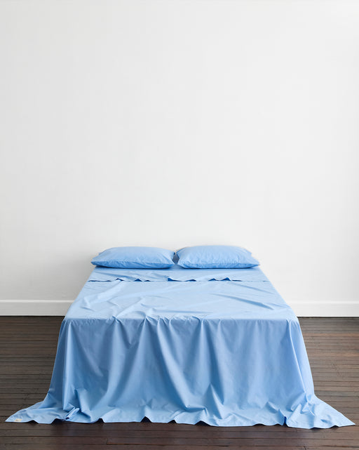 A neatly made bed features the Bed Threads Sky Organic Cotton Flat Sheet and two matching pillows, set against a plain white wall on a dark wooden floor.