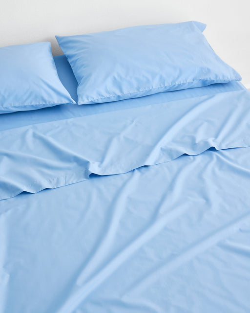 A neatly made bed features the Bed Threads Sky Organic Cotton Flat Sheet, GOTS certified, in light blue with two matching pillows against a white background. Slight wrinkles on the sheet add a relaxed, comfortable look.