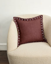 A cream-colored armchair with a square maroon pillow featuring the Bed Threads Cacao & Coast 100% French Flax Linen Swirl Cushion Cover, decorated with white swirl embroidery, sits by a white wall and dark wooden floor. - view