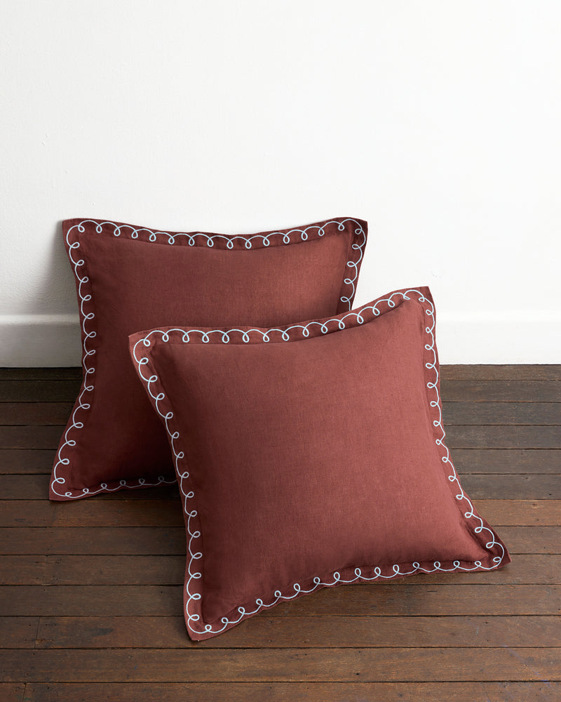 Two square Cacao & Coast 100% French Flax Linen Swirl European Pillowcases by Bed Threads, featuring white decorative edging, rest on a wooden floor against a white wall.