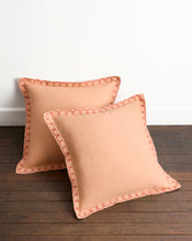 The Bed Threads Terracotta & Pink Clay 100% French Flax Linen Swirl European Pillowcases (Set of Two) are displayed on a wooden floor, leaning against a white wall, highlighting their pink embroidered trim and hidden zip closure. - view