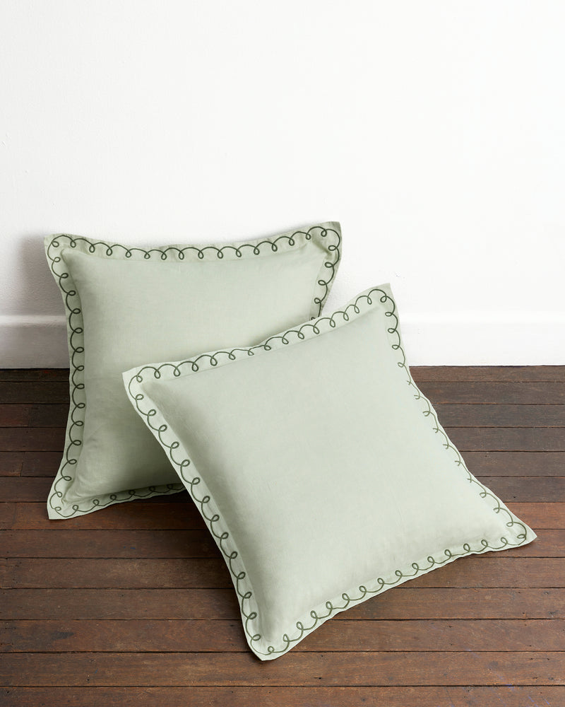 Two Sage & Olive 100% French Flax Linen Swirl European Pillowcases by Bed Threads are arranged on a wooden floor against a white wall—one upright, one flat—showcasing their stylish hue and craftsmanship.
