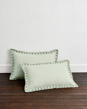 Two Sage & Olive 100% French Flax Linen Swirl Pillowcases by Bed Threads, in light green with decorative dark green stitching, rest on a dark wooden floor against a plain white wall. - view