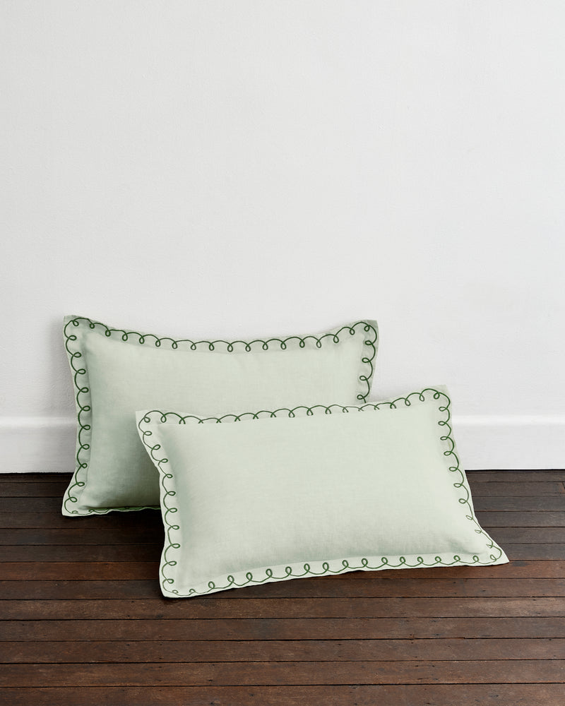 Two Sage & Olive 100% French Flax Linen Swirl Pillowcases by Bed Threads, in light green with decorative dark green stitching, rest on a dark wooden floor against a plain white wall.