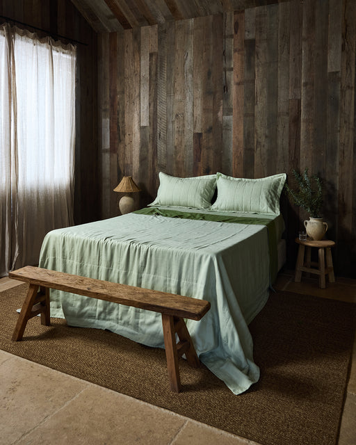 A rustic bedroom with wooden walls, a bed topped with Bed Threads' Sage & Olive Linen Quilt, two pillows, a wooden bench at the foot, a woven rug, and a side table with a ceramic jug and greenery. Sunlight filters through sheer curtains.