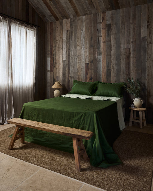 A rustic bedroom with wood-paneled walls, a bed dressed in the Bed Threads Sage & Olive Linen Quilt, a wooden bench at the foot, a woven rug, a small bedside table with a lamp, and sheer curtains letting in natural light.