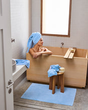 A woman with a blue towel around her head relaxes in a wooden bathtub in a minimalist bathroom. Nearby, plush Bed Threads Sky 100% Cotton Terry Bath Sheets are neatly arranged as natural light pours in. - view