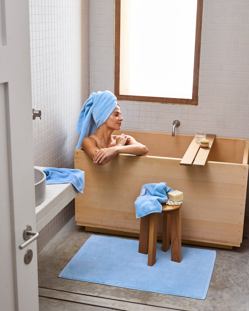 A woman with a blue towel around her head relaxes in a wooden bathtub in a minimalist bathroom. Nearby, plush Bed Threads Sky 100% Cotton Terry Bath Sheets are neatly arranged as natural light pours in.