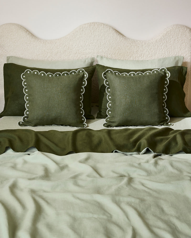 A neatly made bed features a scalloped cream headboard, green French flax linen bedding, and two Bed Threads Olive & Sage 100% French Flax Linen Arch Cushion Covers with white scalloped trim in front of matching shams.