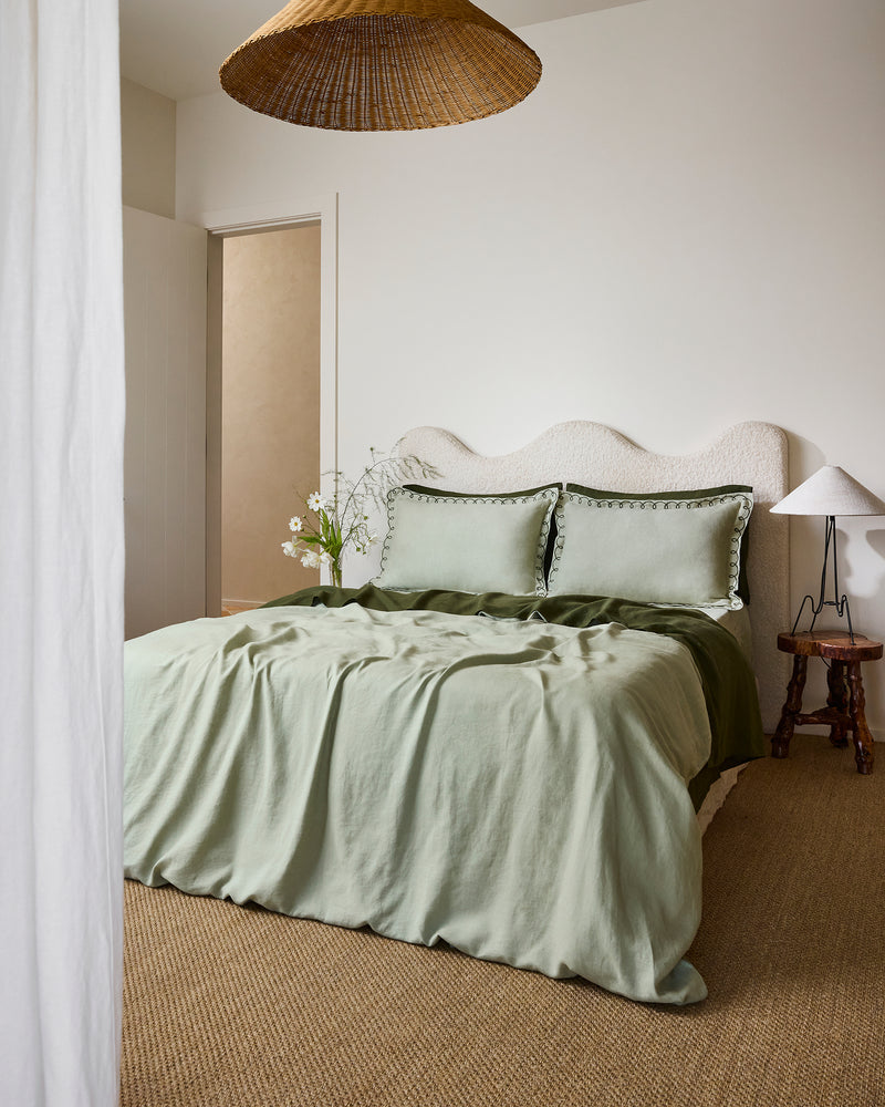 A bed dressed in light green and olive tones features Bed Threads’ Sage & Olive 100% French Flax Linen Swirl Pillowcases, two large swirl-edged pillows, a curved upholstered headboard, and a rustic wooden side table with a white lamp.