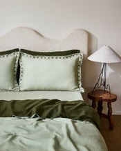 A bed dressed in light green and olive tones features Bed Threads’ Sage & Olive 100% French Flax Linen Swirl Pillowcases, two large swirl-edged pillows, a curved upholstered headboard, and a rustic wooden side table with a white lamp. - view