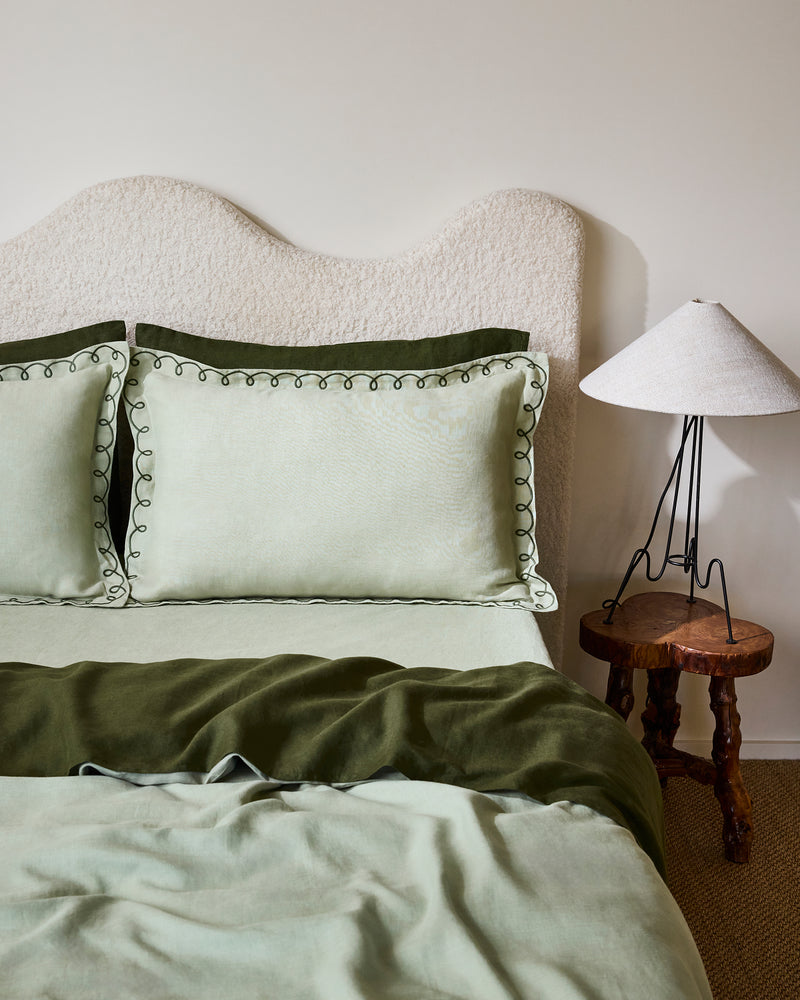 A bed dressed in light green and olive tones features Bed Threads’ Sage & Olive 100% French Flax Linen Swirl Pillowcases, two large swirl-edged pillows, a curved upholstered headboard, and a rustic wooden side table with a white lamp.