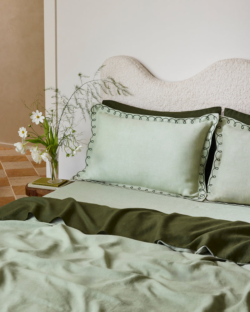 A neatly made bed features Bed Threads’ Sage & Olive 100% French Flax Linen Swirl Pillowcases, an olive green blanket, and a light green duvet. A vase with white flowers rests on the bedside table next to the bed.