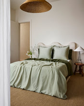 A neatly made bed features Bed Threads’ Sage & Olive 100% French Flax Linen Swirl European Pillowcases (Set of Two), paired with light green bedding, a textured off-white headboard, and a wooden side table holding a glass vase with white wildflowers. - view