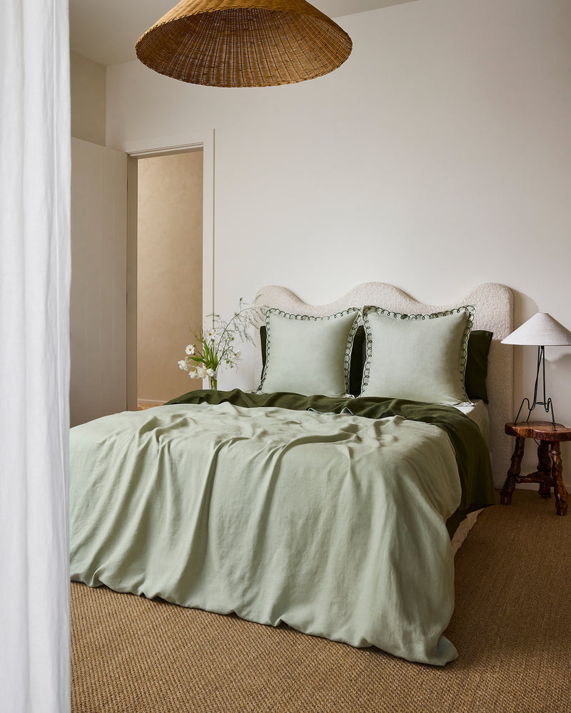 A neatly made bed features Bed Threads’ Sage & Olive 100% French Flax Linen Swirl European Pillowcases (Set of Two), paired with light green bedding, a textured off-white headboard, and a wooden side table holding a glass vase with white wildflowers.