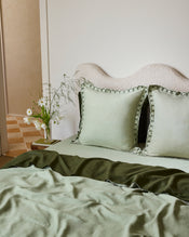 A neatly made bed with sage and olive bedding, two Bed Threads Sage & Olive 100% French Flax Linen Swirl European Pillowcases, and a wavy cream headboard. A small table beside the bed holds a vase of white flowers. - view