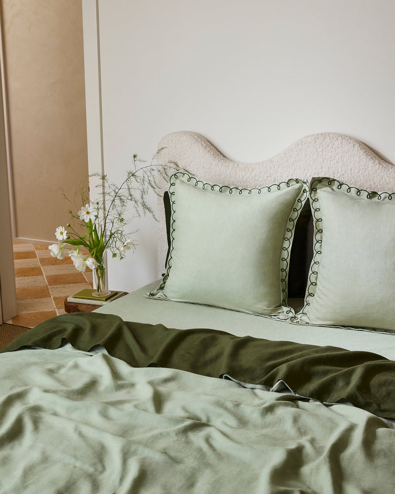 A neatly made bed with sage and olive bedding, two Bed Threads Sage & Olive 100% French Flax Linen Swirl European Pillowcases, and a wavy cream headboard. A small table beside the bed holds a vase of white flowers.