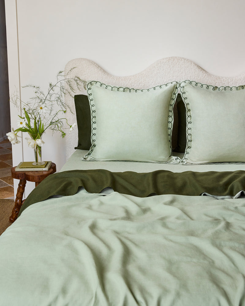A neatly made bed features Bed Threads’ Sage & Olive 100% French Flax Linen Swirl European Pillowcases (Set of Two), paired with light green bedding, a textured off-white headboard, and a wooden side table holding a glass vase with white wildflowers.