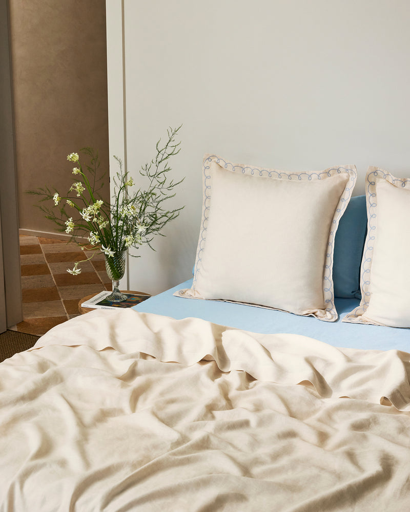A neatly made bed features Bed Threads' Crème & Coast 100% French Flax Linen Swirl European Pillowcases (Set of Two) in cream, set against a light wall and beside a small table with a vase of white flowers and greenery.