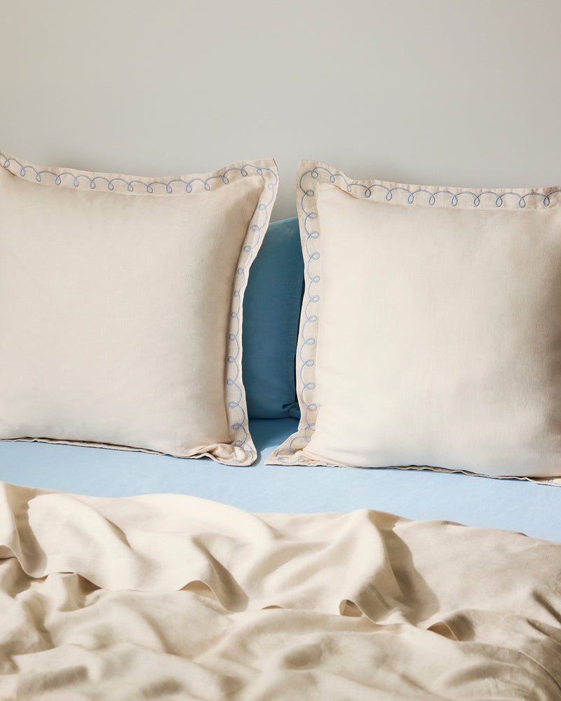 Two Crème & Coast 100% French Flax Linen Swirl European Pillowcases by Bed Threads rest against a light blue headboard, paired with a light beige sheet and soft natural lighting for an inviting look.