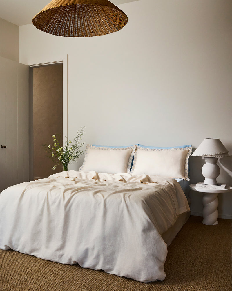 Two Crème & Coast 100% French Flax Linen Swirl Pillowcases by Bed Threads, in cream with blue embroidered edges, rest against a light blue headboard and bedding. The soft, breathable linen appears slightly wrinkled for a relaxed look.