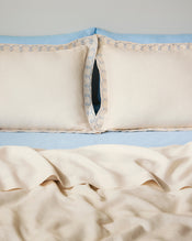 Two Crème & Coast 100% French Flax Linen Swirl Pillowcases by Bed Threads, in cream with blue embroidered edges, rest against a light blue headboard and bedding. The soft, breathable linen appears slightly wrinkled for a relaxed look. - view