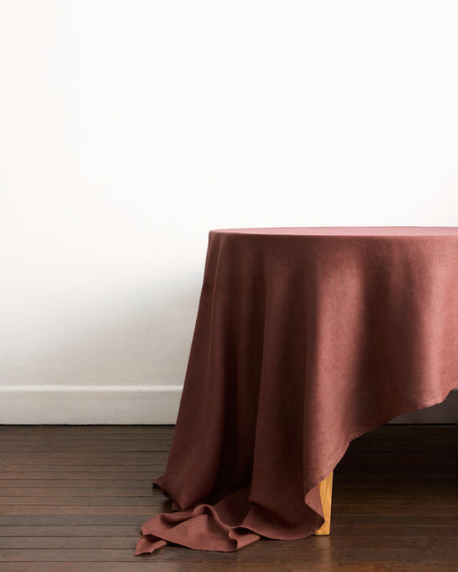 A wooden table, partially covered by the Bed Threads Cacao & Hazelnut 100% French Flax Linen Table Bundle, sits on a dark wooden floor against a plain white wall.