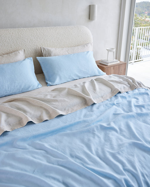 A neatly made bed with Bed Threads Oatmeal 100% French Flax Linen Pillowcases, a beige blanket, and a textured cream headboard sits beside a wooden bedside table and a large window with an inviting outdoor view.