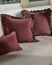 Three Cacao & Coast 100% French Flax Linen Swirl Cushion Covers by Bed Threads—two square, one rectangular with decorative white trim—are arranged on a beige sofa near a window with patterned glass. - view