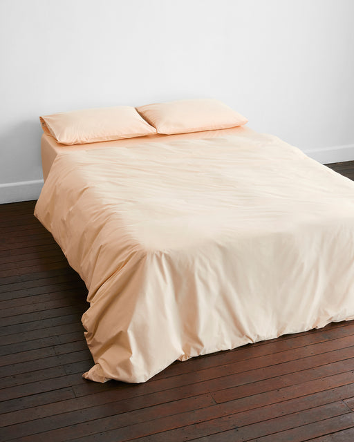 A neatly made bed with the Vanilla Organic Cotton Bedding Set by Bed Threads and two matching pillows rests on a dark wooden floor against a plain white wall.