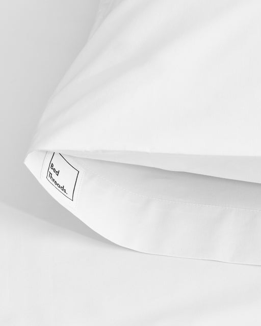 Close-up of Bed Threads’ Crisp White Organic Cotton Pillowcases (Set of Two), featuring a small sewn-in brand label. One pillowcase is partially tucked in, set against a smooth white background.