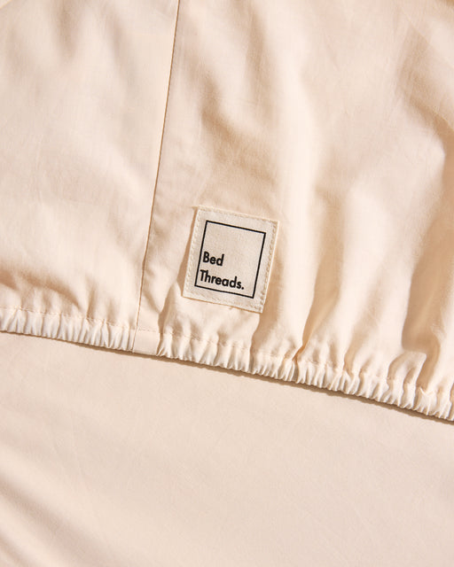 A close-up shows a “Bed Threads” label on the Vanilla Organic Cotton Fitted Sheet. The cream-colored percale fabric appears smooth and slightly gathered along a stitched seam, highlighting its all-natural, organic cotton material.