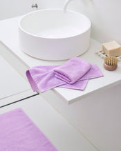 A modern white bathroom sink with a stack of Bed Threads’ Violet 100% Cotton Terry Face Washers (Set of Two), plus a beige soap bar, wooden brush, and a matching violet bath mat on the floor. - view
