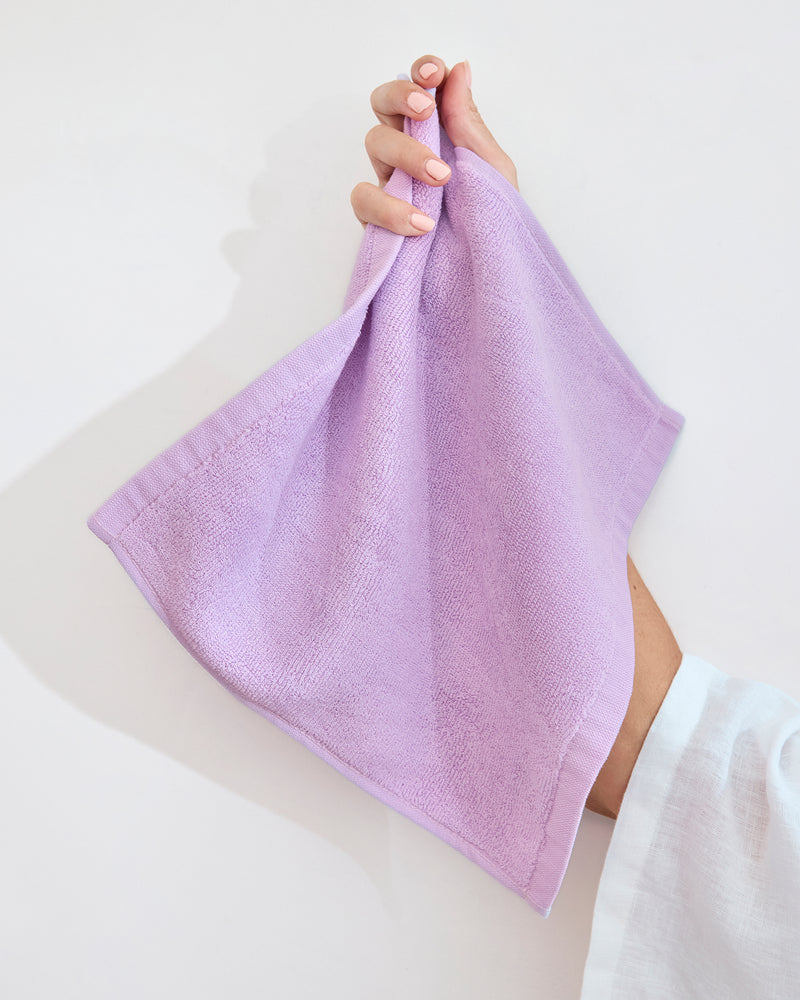 A modern white bathroom sink with a stack of Bed Threads’ Violet 100% Cotton Terry Face Washers (Set of Two), plus a beige soap bar, wooden brush, and a matching violet bath mat on the floor.