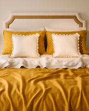 A neatly made bed with a beige upholstered headboard, two mustard yellow pillows, and two white Bed Threads Crème & Turmeric 100% French Flax Linen Arch Cushion Covers, layered with a mustard yellow bedspread over crisp linen sheets. - view