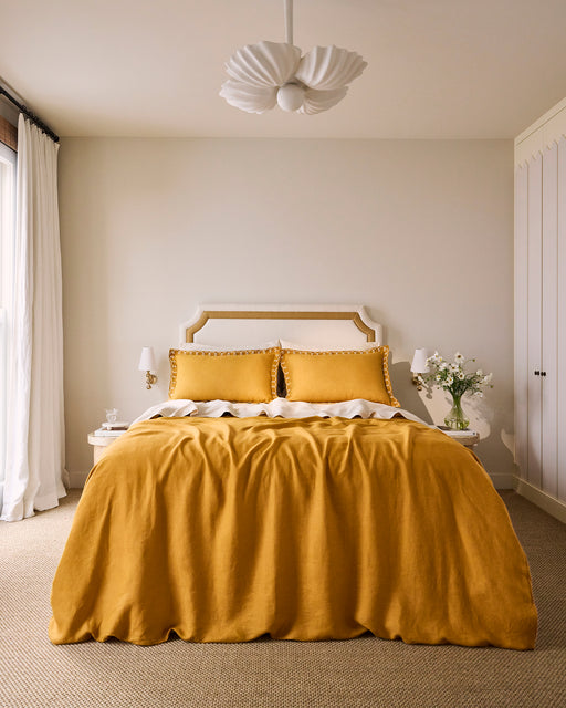 A neatly made bed with a beige and gold headboard, white sheets, a mustard yellow blanket, and Bed Threads Turmeric & Crème 100% French Flax Linen Swirl Pillowcases (Set of Two) in mustard yellow.