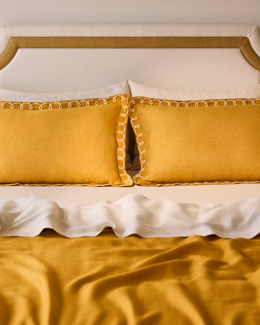 A neatly made bed with a beige and gold headboard, white sheets, a mustard yellow blanket, and Bed Threads Turmeric & Crème 100% French Flax Linen Swirl Pillowcases (Set of Two) in mustard yellow.