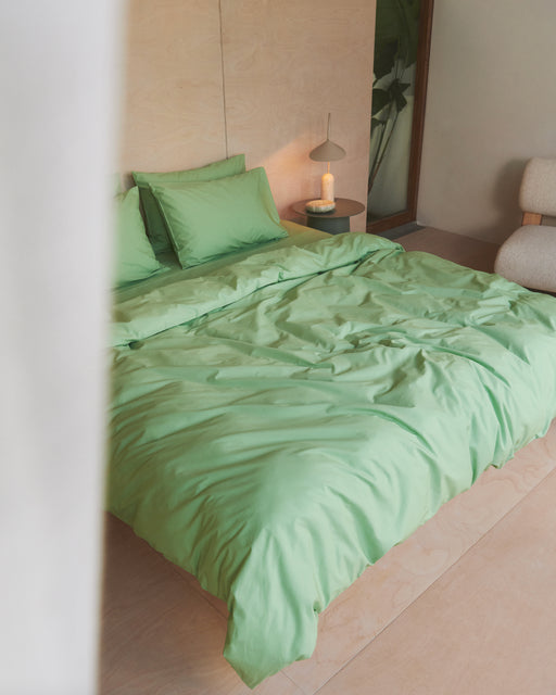 A neatly made bed with the Bed Threads Apple Organic Cotton Duvet Cover sits in a minimal, modern bedroom with light wood floors and walls, a small round nightstand, a lamp, and a lit candle.