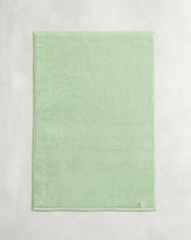 The Bed Threads Apple 100% Cotton Terry Bath Mat, in light green, is neatly folded and laid flat on a light gray surface with a small white label sewn onto one corner. - view