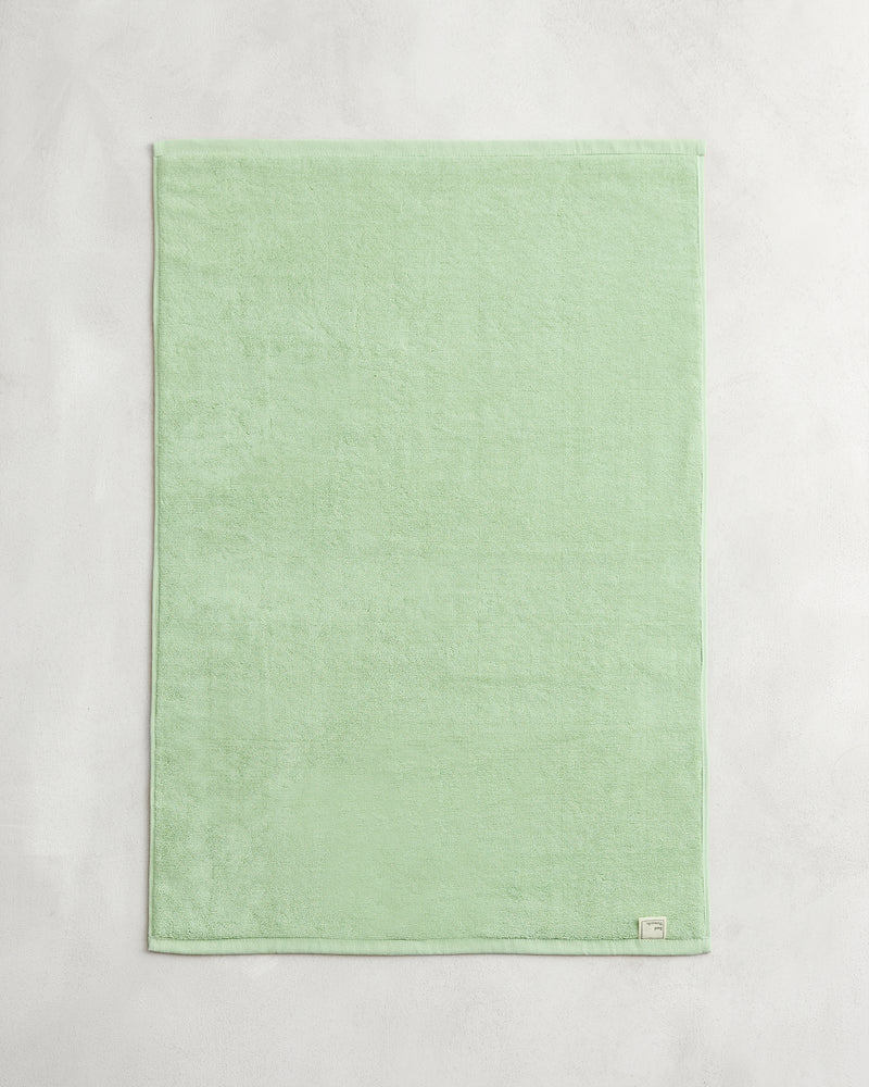 The Bed Threads Apple 100% Cotton Terry Bath Mat, in light green, is neatly folded and laid flat on a light gray surface with a small white label sewn onto one corner.