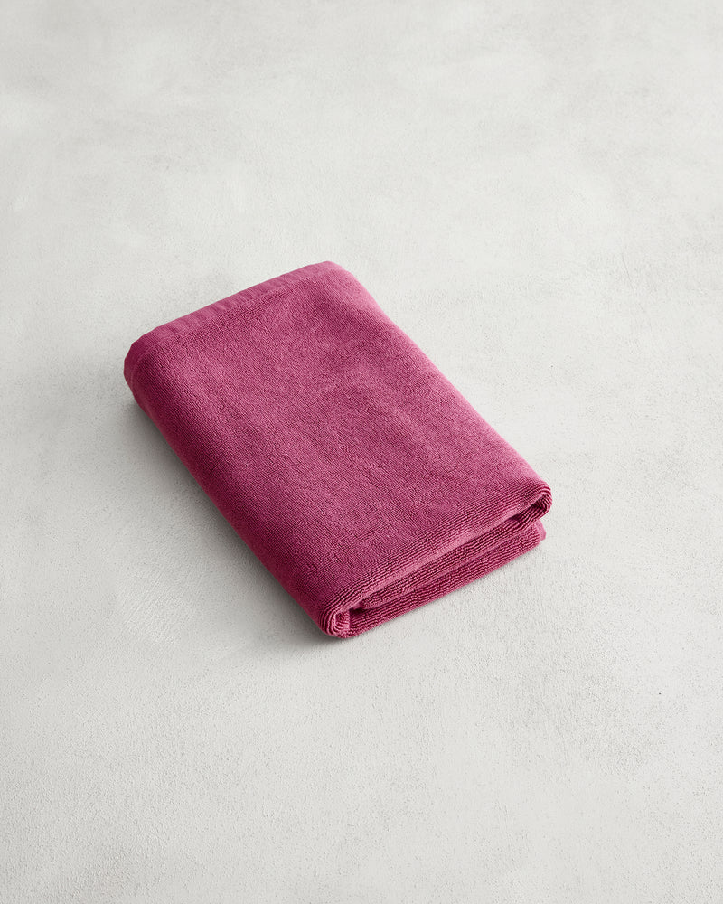 A plush Berry 100% Cotton Terry Bath Mat by Bed Threads lies on a light gray, textured surface next to a neatly folded, soft magenta towel.