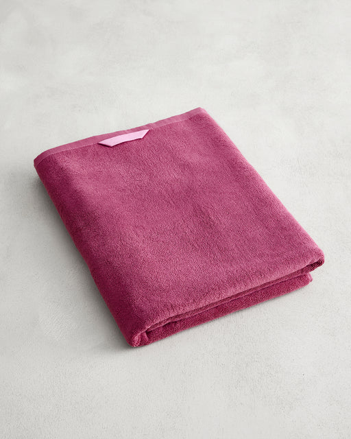 The Bed Threads Berry 100% Cotton Terry Bath Sheet, in plush raspberry, is neatly folded on a light gray surface with a light pink hanging loop visible. Quick-drying and stylishly functional, it's the perfect towel for any bathroom.