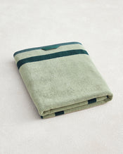 The Bed Threads Forest & Sage Two-Toned 100% Cotton Terry Bath Sheet features a folded light green design with two dark green stripes near the edges, crafted from soft cotton and displayed on a light textured surface. - view